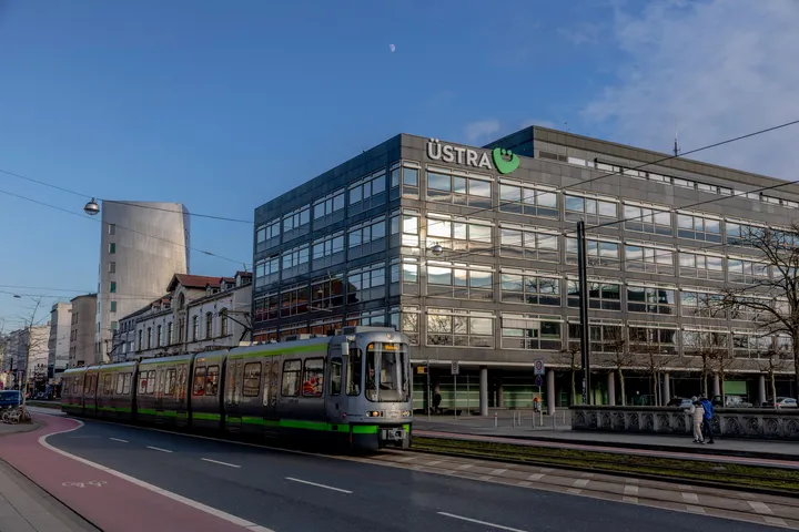 Eine silberne Stadtbahn fährt an der Zentrale der ÜSTRA am Hohen Ufer vorbei. Die Sonne geht gerade unter und taucht das Gebäude in ein warmes Licht.