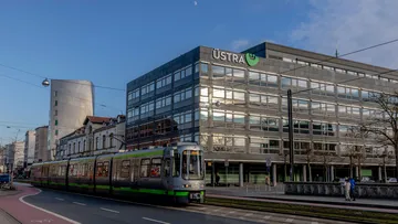 Eine silberne Stadtbahn fährt an der Zentrale der ÜSTRA am Hohen Ufer vorbei. Die Sonne geht gerade unter und taucht das Gebäude in ein warmes Licht.