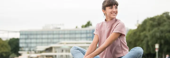 Eine junge Frau sitzt im Schneidersitz am Maschsee und lacht, im Hintergrund ist ein Gebäudekomplex sichtbar