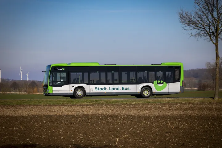 Ein ÜSTRA Bus fährt auf einer Landstraße zwischen Feldern. Auf dem Bus ist das ÜMO Logo zu sehen und die Aufschrift "Stadt. Land. Bus". Im Hintergrund sind Felder, Bäume und Windräder zu sehen.