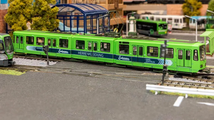 Das TW 6002 Modell fährt auf Schienen in einer Modellbaulandschaft. Auf seiner Seite ist eine Beklebung: "Herrenbekleidung Erdmann". Im Vordergrund sieht man Straße. Der Hintergrund ist unscharf. Dort stehen viele Modellbusse, Gebäude und Bäume.