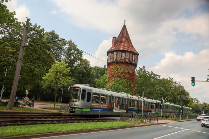 Im Hintergrund sieht man den Döhrener Turm in Hannover, aus roten Backsteinen mit spitzem Dach. Vor ihm fährt eine silberne Stadtbahn, ein TW2000 von rechts nach links durchs Bild. Im Vordergrund ist eine Straße zu sehen, im Hintergrund Bäume, etwas blauer Himmel und weiße und graue Wolken.