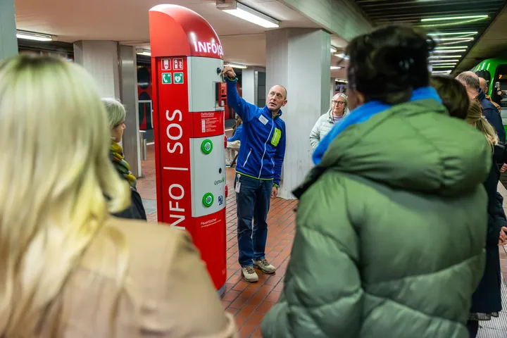 Eine Gruppe Menschen befinden sich in der Station Hauptbahnhof und nehmen an einer Führung zum Thema Sicherheit teil. Sie stehen im dem Rücken zum Betrachter des Bildes und schauen auf eine Notrufsäule und einen Mann, der diese erklärt.