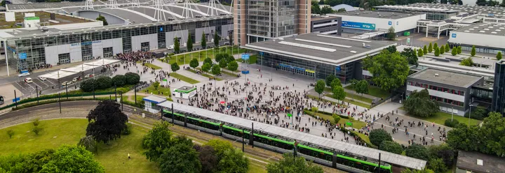 Eine Drohnenaufnahme zeigt eine ÜSTRA Stadtbahn an der Messe. Im Hintergrund sind viele Menschen, Bäume, das Messegelände und Gebäude zu sehen.
