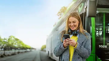 Frau steht vor einem Bus und schaut auf ihr Handy, dabei lacht sie
