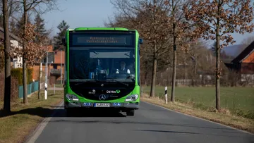 Ein ÜSTRA Bus fährt über eine Landstraße. Zu sehen ist der Bus von vorne. Im Hintergrund sind Bäume, Häuser und Felder zu sehen.