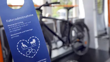 A bicycle is standing in the multi-purpose area of a light rail vehicle, blurred in the background. In the foreground is a blue flyer with the headline “Bicycle transport” and the times when this is permitted.