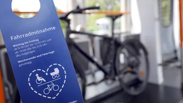 A bicycle is standing in the multi-purpose area of a light rail vehicle, blurred in the background. In the foreground is a blue flyer with the headline “Bicycle transport” and the times when this is permitted.