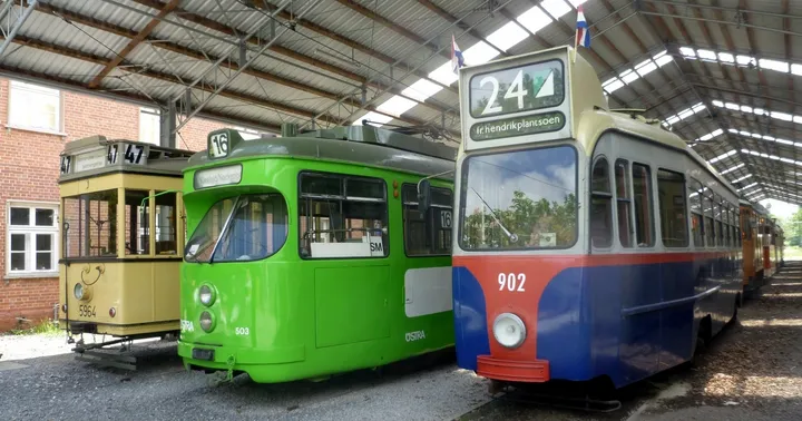 Drei alte bunte Stadtbahnen nebeneinander in einer Halle. Die linke Bahn ist gelb, die mittlere knallgrün und die rechte blau-rot.