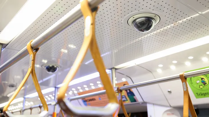 Überwachungskamera an der Decke eines öffentlichen Verkehrsmittels. Zu sehen sind auch Halteschlaufen, die an einer Stange hängen.