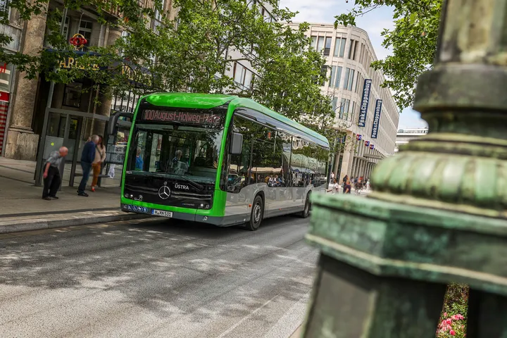 Ein Bus der Linie 100 steht an der Haltestelle Kröpcke vor dem GOP in Hannovers Innenstadt. Der Fotograf steht auf der gegenüberliegenden Straßenseite und blickt hinter einer Steinsäule hervor.