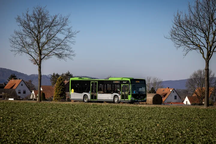 Ein ÜSTRA Bus fährt auf einer Landstraße. Im Hintergrund sind Bäume, Häuser und Berge zu sehen.