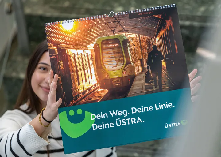 Eine junge Frau hält den ÜSTRA Bildkalender 2026 in die Höhe. Ihr Gesicht ist dabei zur Hälfte bedeckt.