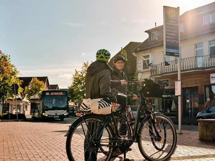 Two people are standing with their bicycles at a bus stop in Steinhude. They are wearing helmets and waiting for the bus, which is just pulling up. The sun is shining, the sky is blue.