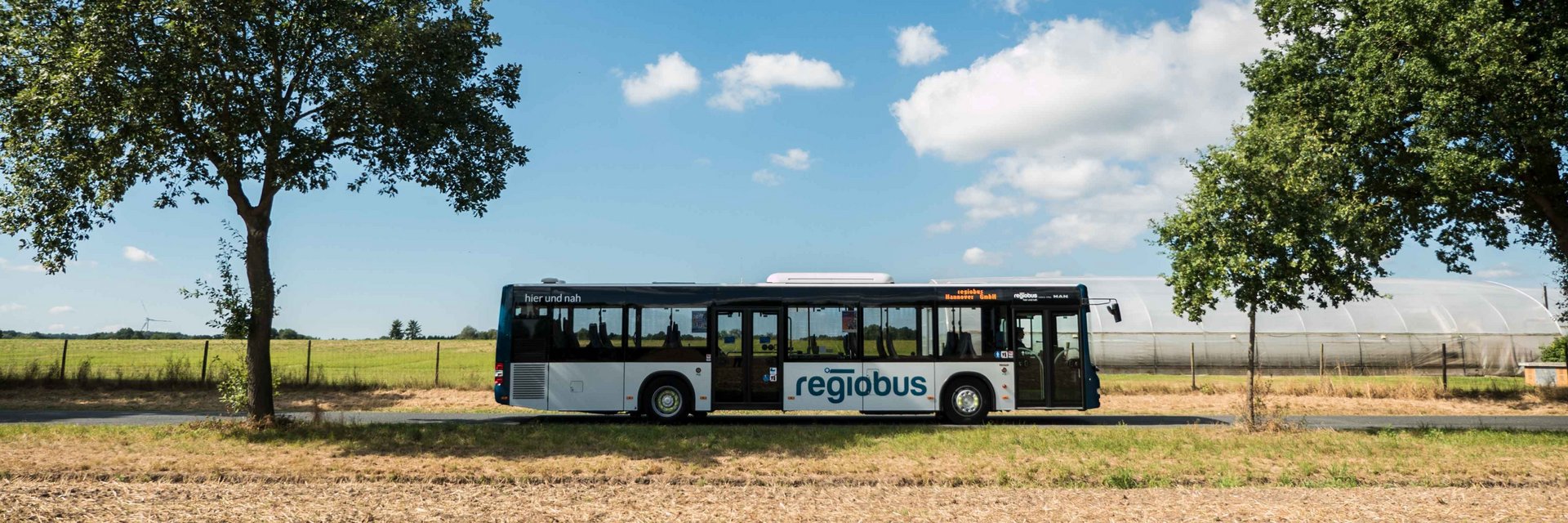Ein Bus der regiobus GmbH fährt auf einer Landtsraße entlang. Es ist blauer Himmel im Hintergrund und ein Stoppelfeld im Vordergrund zu sehen.